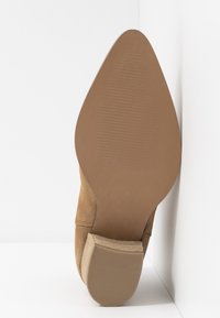 Beżowy zamszowy botek za kostkę z ostro zakończonym noskiem i niskim, szerokim obcasem. Podeszwa jest gładka z subtelną żebrowaną fakturą dla zapewnienia przyczepności.