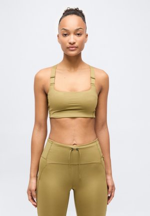 Kobieta stojąca przodem, ubrana w oliwkowo zielony sportowy stanik i legginsy z wysokim stanem na jasnym, prostym tle.