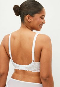 Reggiseno bianco in pizzo con spalline regolabili, caratterizzato da un motivo floreale e una chiusura a gancio sul retro. Texture liscia e delicata.