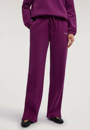 Personne portant un pantalon large violet avec cordon de serrage et un haut à manches longues assorti, associé à des chaussures noires pointues, debout devant un fond uni.