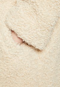 Tessuto in pile color crema con una superficie morbida e texturizzata, dotato di una tasca a mano visibile. Il materiale appare caldo e accogliente.