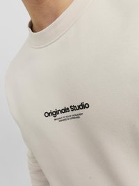 Lichtbeige sweater met een ronde hals en een zachte textuur. Heeft een zwart "Originals Studio" logo en extra witte tekst eronder.
