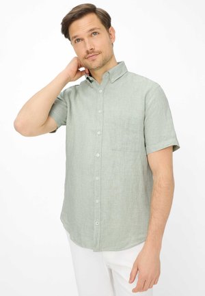 Mann trägt ein hellgrünes, kurzärmeliges Hemd mit Knöpfen und Tasche, weiße Hose, berührt den Hals und blickt leicht nach oben vor weißem Hintergrund.