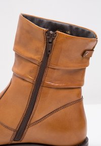 Gros plan d'une botte cheville en cuir beige avec fermeture éclair noire, revers plié et détails cousus sur un fond clair uni.