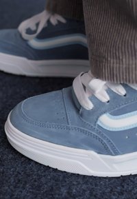 Plave suede tenisice s bijelim vezicama i bočnim naglascima. Imaju teksturiranu poslovnicu i perforacije na prstima, što omogućava ventilaciju.