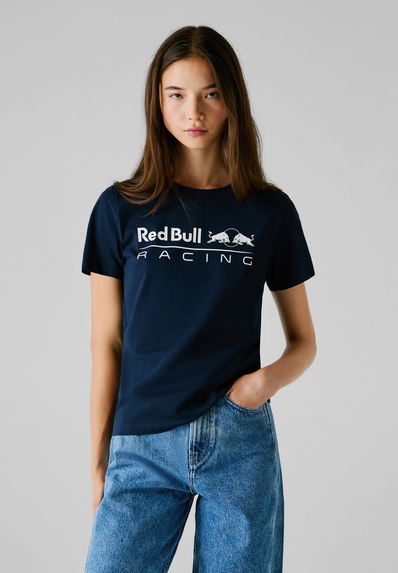 T-shirt in cotone blu navy con maniche corte, caratterizzato dal logo "Red Bull Racing" in bianco. Abbinato a jeans in denim blu a vita alta.