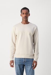 Sudadera de un tono beige claro con cuello redondo, con el logotipo "BOSS" en relieve en la parte delantera, y puños y bajo acanalados. Combinada con jeans azules.