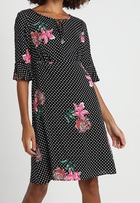Musta pilkullinen mekko vaaleanpunaisilla kukkakuoseilla, lyhyet hihat ja pyöreä pääntie. Mekossa on solmitys yksityiskohtana kaulassa ja rento istuvuus.