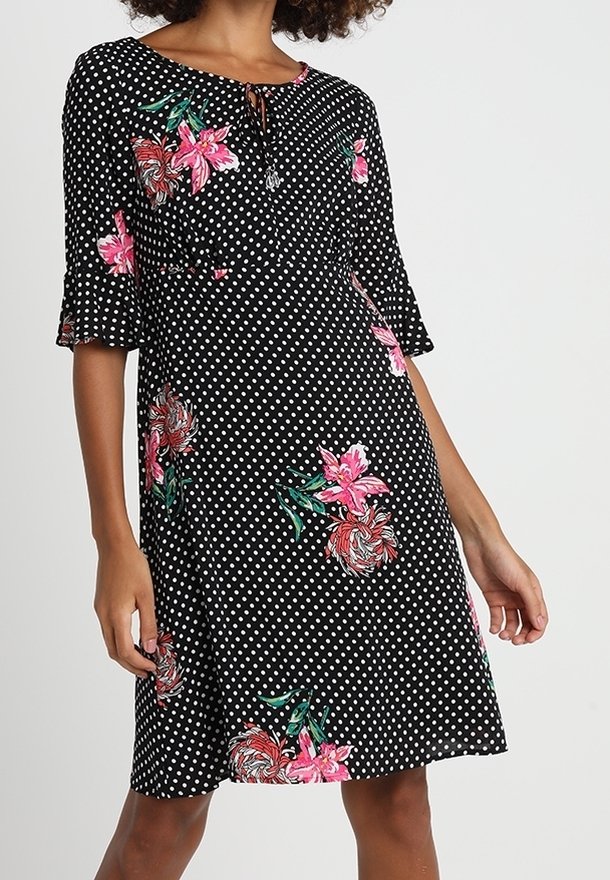 Musta pilkullinen mekko vaaleanpunaisilla kukkakuoseilla, lyhyet hihat ja pyöreä pääntie. Mekossa on solmitys yksityiskohtana kaulassa ja rento istuvuus.