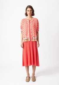Korallfärgad kofta med geometriskt mönster och strukturerade ärmar, i kombination med en matchande veckad korallklänning. Modellen står i neutrala skor.