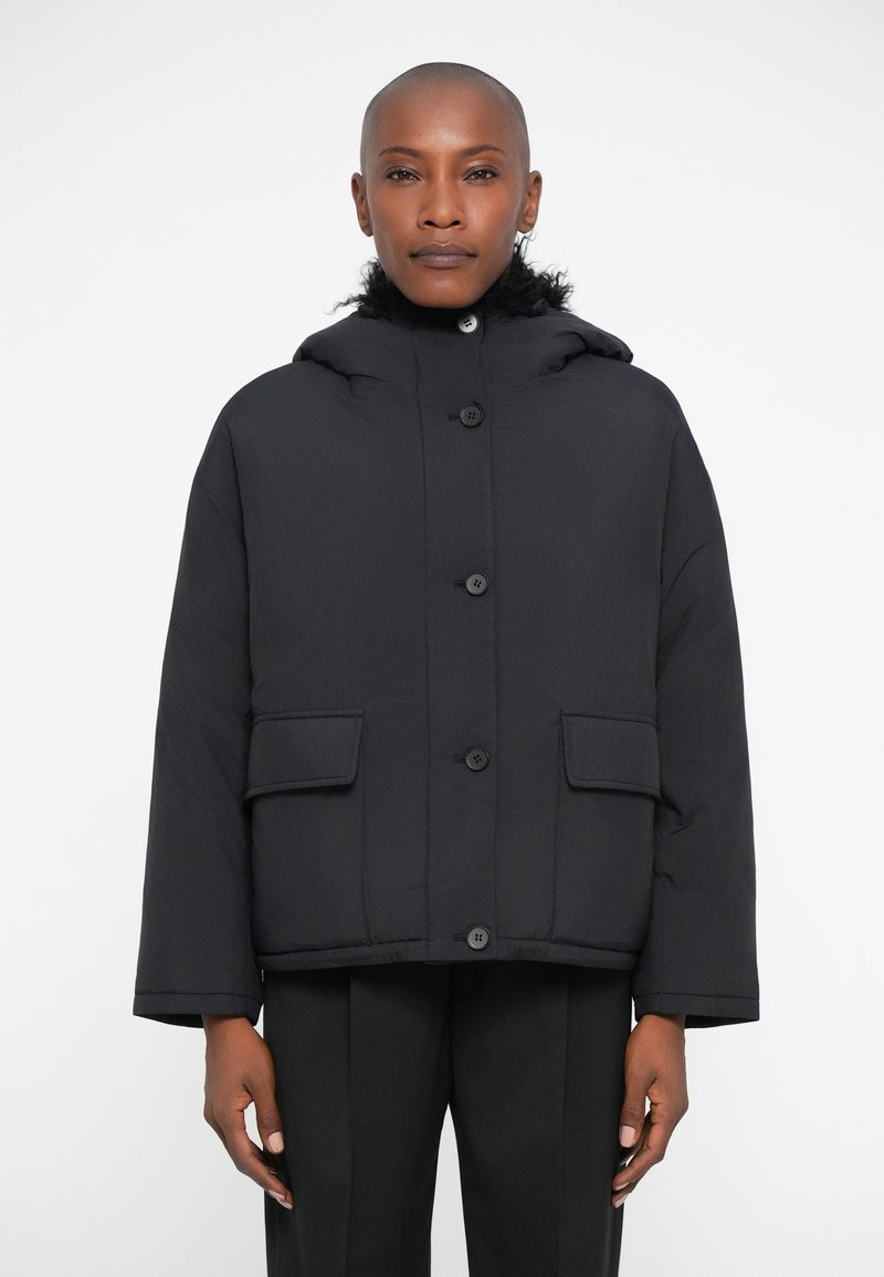 Giacca a vento nera con cappuccio, caratterizzata da grandi bottoni frontali e due tasche anteriori. Tessuto liscio con un design cropped e una vestibilità rilassata.