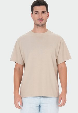 Mann mit kurzen dunklen Haaren, der ein schlichtes beiges T-Shirt mit kurzen Ärmeln und hellblaue Jeans trägt, steht vor einem weißen Hintergrund.