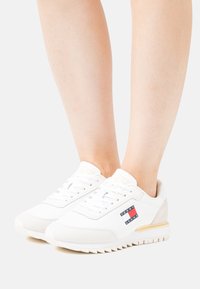 Bílé sportovní boty s texturovaným textilním svrškem, světle béžovými akcenty a výrazným logem Tommy Jeans. Disponují polstrovanou podrážkou se žebrováním.