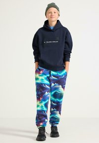 Granatowa bluza z kapturem z logo z przodu, kolorowe spodnie dresowe w wzory z odcieniami niebieskiego, zielonego i fioletowego oraz czarne buty sportowe.