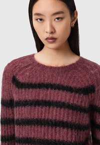 Jeune femme aux cheveux raides et foncés, portant un pull violet duveteux à rayures horizontales noires et une petite boucle d'oreille en or.