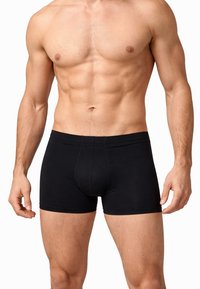 Uomo muscoloso che indossa boxer neri, in piedi con le braccia rilassate lungo i fianchi su uno sfondo bianco.