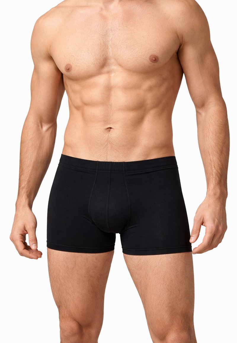Uomo muscoloso che indossa boxer neri, in piedi con le braccia rilassate lungo i fianchi su uno sfondo bianco.