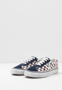 Matala sneakers dengan ujung suede navy, kanvas kotak-kotak merah dan hitam, tali sepatu putih, dan sol karet putih di latar belakang putih.