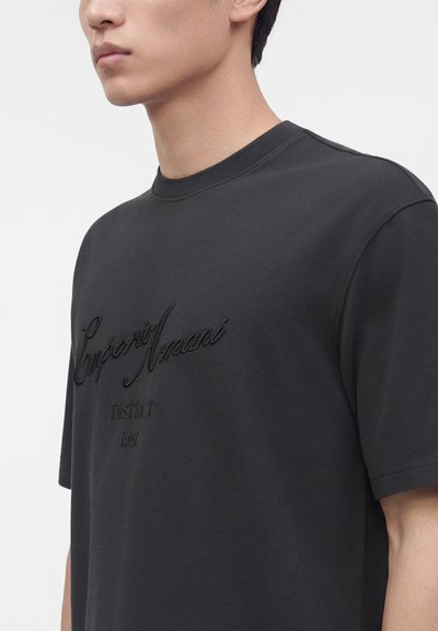 Νεαρός άνδρας που φοράει μαύρο t-shirt Emporio Armani με στρογγυλή λαιμόκοψη, με ανάγλυφο λογότυπο και κείμενο στο στήθος, εμφανιζόμενος από τους ώμους και πάνω.
