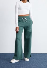 Žena ve zelených širokých teplácích Nike s páskem na stažení, bílém dlouhém krátkém topu a bílých teniskách stojící na modrém koberci.