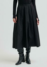 Osoba ubrana w czarną bluzkę z długim rękawem, czarną midi spódnicę z wysokim stanem z plisami oraz czarne skórzane botki na obcasie na jednolitym tle.