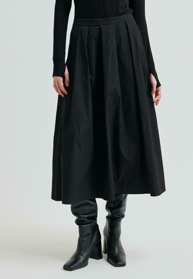 Osoba ubrana w czarną bluzkę z długim rękawem, czarną midi spódnicę z wysokim stanem z plisami oraz czarne skórzane botki na obcasie na jednolitym tle.