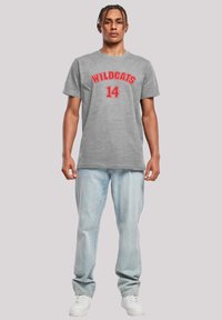 Pilkos spalvos medvilninis marškinėliai su užrašu "WILDCATS" ir numeriu "14" raudonais simboliais, derinami su šviesiai mėlynomis džinsinėmis kelnėmis ir baltomis sportinėmis bateliais.