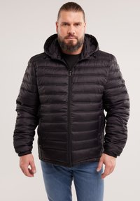 Hombre con barba que lleva una chaqueta acolchada negra con capucha y vaqueros azules, de pie contra un fondo claro y liso.