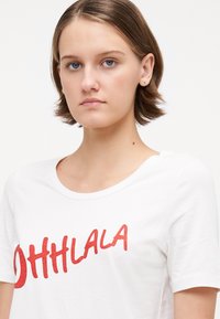 Hvit bomull t-skjorte med rød glittertekst "OHHLA". Den har rund hals og korte ermer med en avslappet passform.