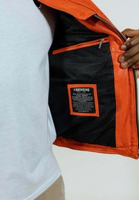 Veste en cuir orange avec une doublure en maille noire, dotée d'une poche intérieure rouge et d'une étiquette de marque avec des instructions d'entretien. Fermeture zippée.