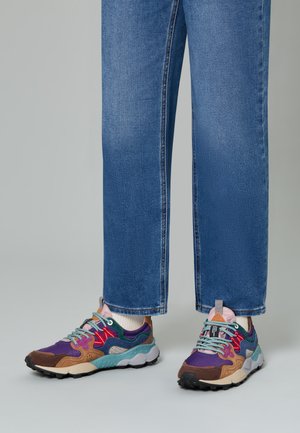Feet in blue jeans wearing colorful sneakers with purple, brown, red, teal, and pink accents, standing on a gray surface.