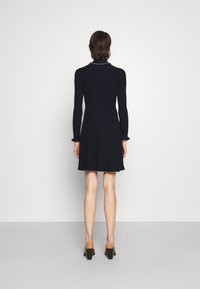 Stickad klänning i marinblått med långa ärmar, ribbat mönster, utsvängd kjol och scalloped krage och ärmkanter; matchad med klackskor.