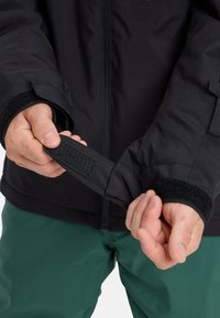 Schwarze Jacke mit verstellbarem Handgelenkabschluss und Klettverschluss. Das Innere hat eine glatte Textur und wird über grünen Hosen getragen.