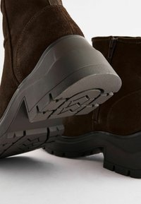 Bottines en daim marron avec une épaisse semelle en caoutchouc noir dotée d'un motif de crans texturé. Comprend un détail de fermeture éclair sur le côté.