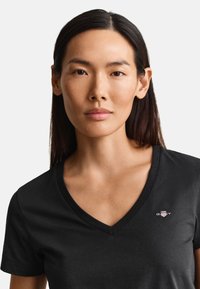Femme aux longs cheveux foncés portant un t-shirt noir col en V avec un petit logo GANT sur la poitrine, face à l’avant avec une expression neutre.