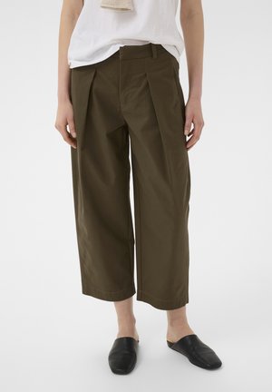 Asmuo dėvi plačias, trumpintas alyvuogių žalios spalvos kelnes, baltą viršutinę dalį ir juodus slip-on batus, stovintis ant paprasto balto fono.