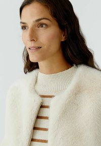 Kunstbont ritjacket in crème, met daaronder een geribde beige trui met horizontale bruine strepen. Zachte textuur zichtbaar.