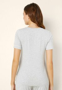 Femeie cu păr lung și brunet îmbracă un tricou simplu cu mânecă scurtă de culoare gri deschis și pantaloni asortați, întorsă cu spatele într-un fundal bej.