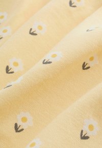 Gult tyg med en subtil struktur, tryckt med små vita blommor och grågröna blad, vilket ger ett mjukt och lätt utseende.