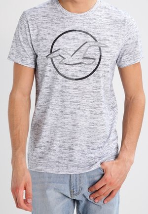 Homme portant un t-shirt gris clair à manches courtes avec une mouette dessinée en noir dans un cercle sur le devant et un jean bleu clair.