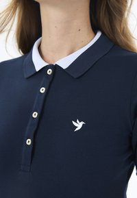 Polo marine avec un col blanc, doté de boutons et d'un petit logo d'oiseau blanc sur la poitrine. Détail de tissu texturé visible.