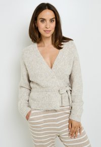 Cardigan avvolgente color beige chiaro con un dettaglio legato, polsini a coste e texture morbida, abbinato a pantaloni rigati beige e bianchi.