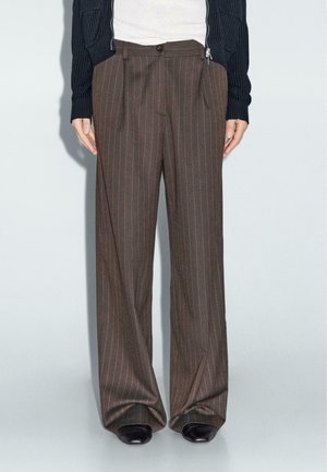 Personne portant un pantalon large marron à fines rayures, des chaussures noires, un haut blanc et un cardigan en maille bleu marine avec les manches baissées.