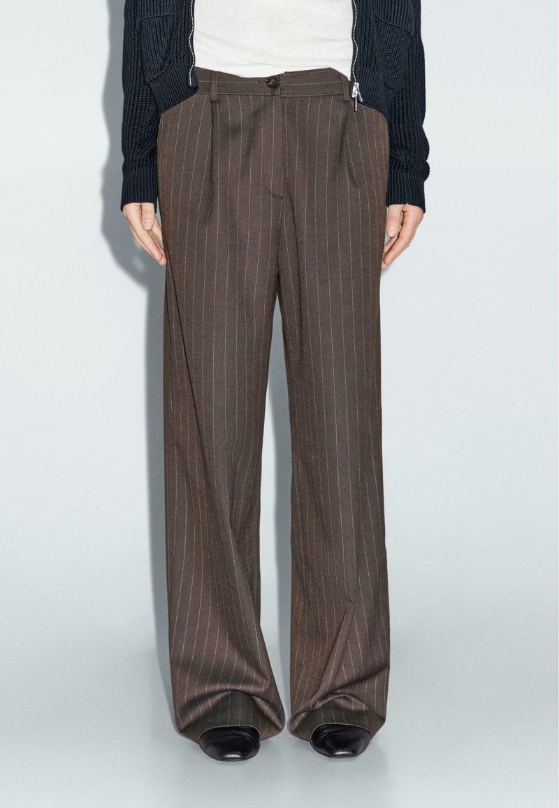 Personne portant un pantalon large marron à fines rayures, des chaussures noires, un haut blanc et un cardigan en maille bleu marine avec les manches baissées.