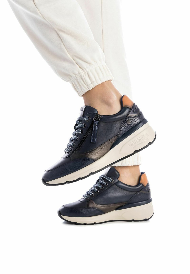 Zapatillas de cuero azul marino con acentos texturizados, que presentan una cremallera, cordones redondos y una suela color crema. Un detalle en marrón claro en la parte trasera añade contraste.