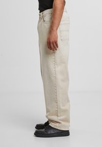 Beige denim jeans displayed from the side, featuring a straight cut, back pocket, and stitching details, paired with black sneakers.