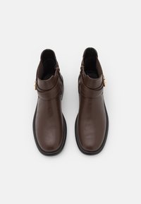 Bottes basses en cuir marron à texture lisse, semelle plate noire, fermetures éclair latérales et une sangle décorative autour de la cheville.