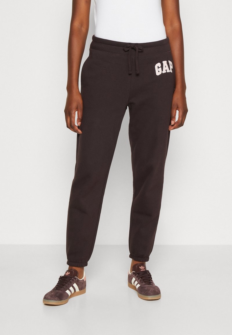 Pantalones de chándal negros con cintura elástica, detalle de cordón y logotipo frontal. Piernas ajustadas y puños de punto. Llevados con zapatillas oscuras.