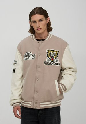 Giacca varsity beige con maniche crema, caratterizzata da ricami di una tigre e dettagli testuali. Polsini e orlo a coste con accenti a strisce.