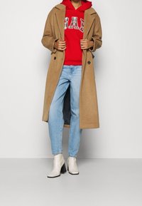 Abrigo de lana camel con solapas sobre una sudadera con capucha roja con un logo a cuadros, combinado con jeans azul claro y botines blancos con tacón.